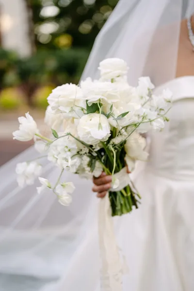 Bridal bouquet of white wedding bouquet blooms with greenery, ribbon-wrapped handle and long ribbons, held against veil and trees outdoors