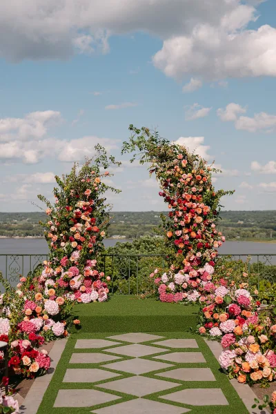 Ceremony aisle decor with a floral arch wedding of roses and hydrangeas, turf runner over stone pavers on a lakeside terrace under clouds