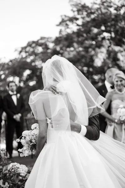 Wedding kiss as the bride and groom share a ceremony kiss moment under a flowing veil, with trees and guests softly blurred behind
