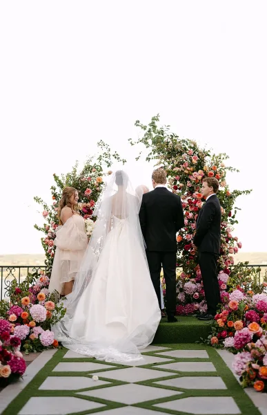 Wedding ceremony with bride in long veil walking to groom and officiant under a rose and hydrangea floral arch on an outdoor terrace