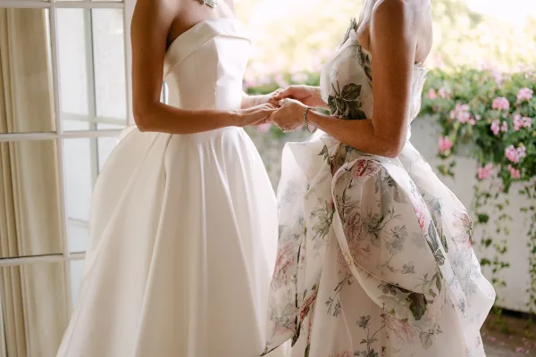Wedding dress fitting as mother in a floral dress adjusts the strapless bridal gown by bright window panes with garden flowers behind