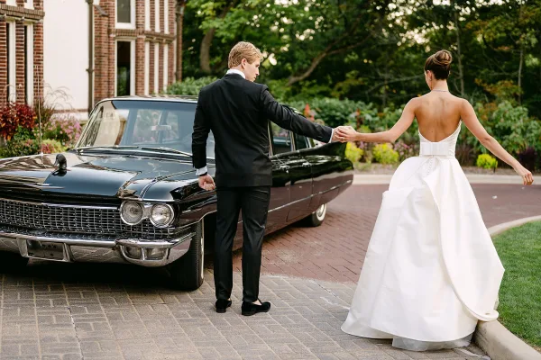 Couple portrait of bride and groom holding hands beside a classic car, bride in a backless satin gown on a brick driveway near a garden estate