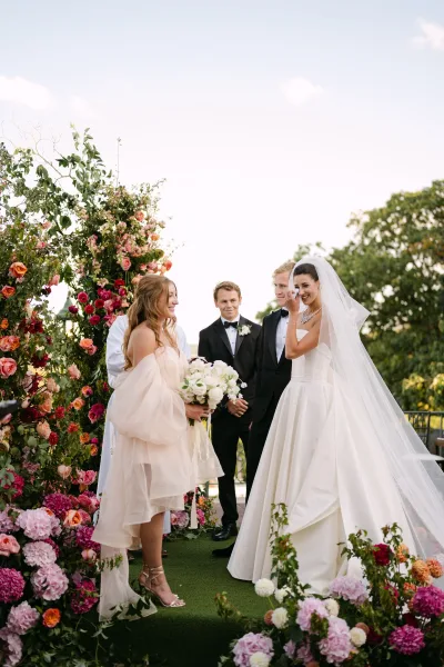 Ceremony moment as bride wipes tears beside groom at a colorful outdoor wedding ceremony under a floral arch in a garden setting