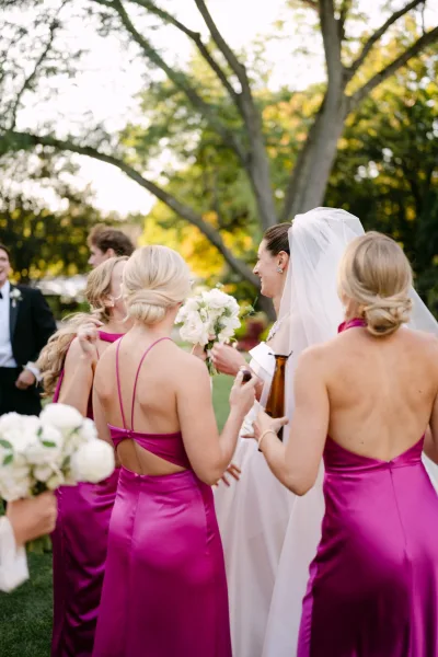 Bridesmaids with bride laughing together in magenta dresses, holding bouquets beside her veil in a lush outdoor garden setting