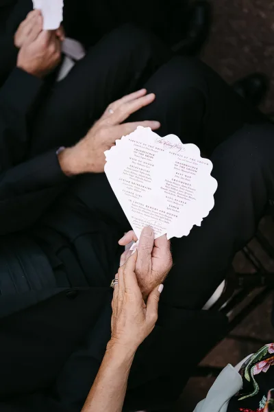 Wedding ceremony program held like a fan by a guest, scalloped paper edge in focus with rings and suit beside ceremony seating