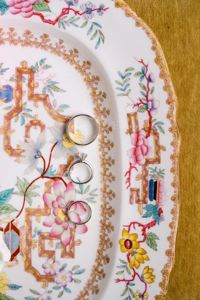 Wedding rings and engagement ring flat lay with diamond solitaire and bands on a floral porcelain plate atop gold fabric background