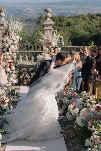 Ceremony kiss moment as the couple share a recessional kiss beneath a long veil on a stone terrace, framed by garden trees and distant hills.