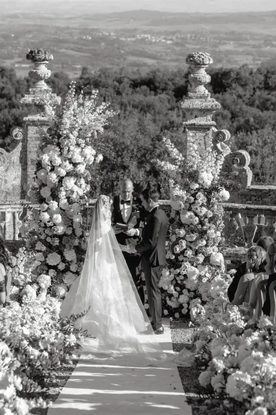 Ceremony moment as the couple are exchanging vows outdoors beneath floral pillars, with a stone balustrade and rolling hills beyond.