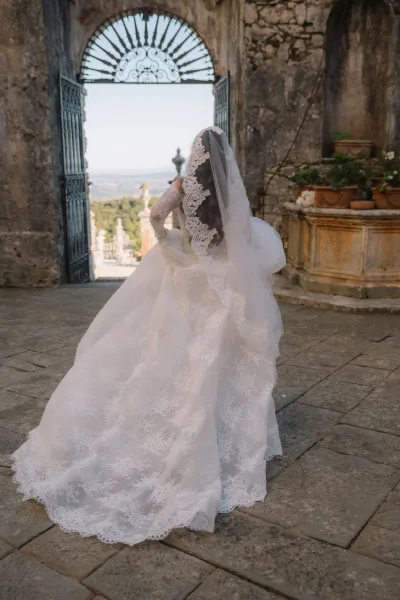 Bridal portrait of a bride with lace veil and a long train, framed by a stone archway and wrought iron gate in a stone courtyard.