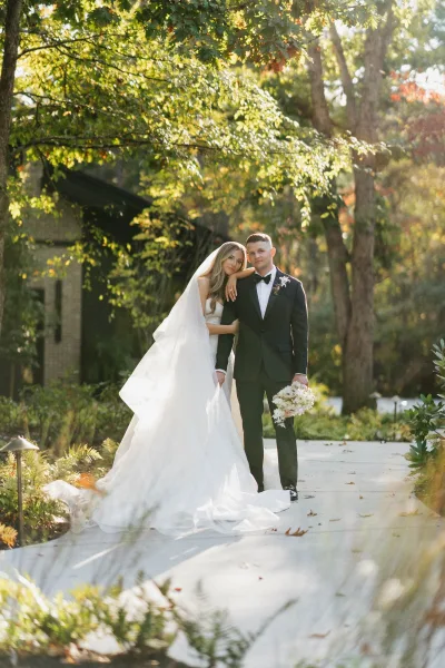 Couple portrait of bride leaning on groom in a sunlit outdoor wedding portrait, cathedral veil and white bouquet on a garden walkway near brick building