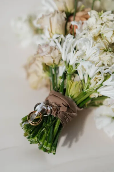 Wedding rings resting on a white bouquet with ribbon wrap on a light neutral surface, featuring gold and silver wedding bands in soft light