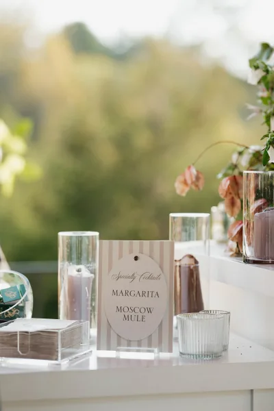 Cocktail menu sign in an acrylic holder on a wedding bar with pillar candles, glass vases, florals, and a mountain-view terrace backdrop