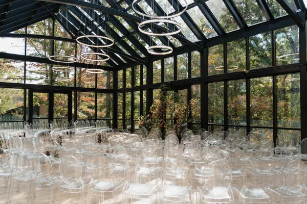 Ceremony setup with indoor ceremony seating, clear acrylic chairs facing a floral installation under ring chandeliers in a glass conservatory with autumn trees outside