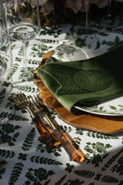 Reception tablescape with green wedding napkins on patterned plates and wooden chargers, amber-handled gold flatware, and mixed glassware in soft shadows