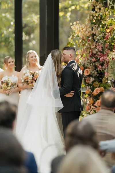 Ceremony kiss as bride and groom embrace beneath a rose and greenery floral arch, her veil flowing in a sunlit glasshouse with guests behind