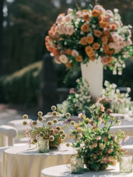 Cocktail table decor arranged as romantic wedding centerpieces on ivory table linens amid sunlit garden greenery and trees.