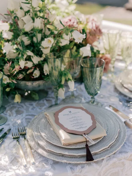 Wedding place setting with layered charger plates on a lace tablecloth, set amid garden greenery on a sunlit outdoor lawn.