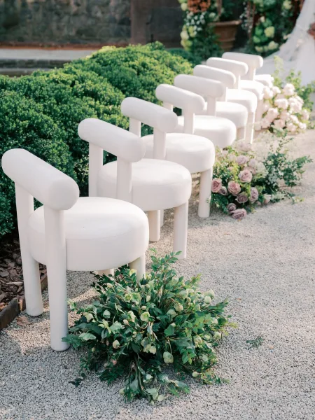 Ceremony seating with modern wedding chairs lining a gravel aisle, edged with roses, set against garden hedges and a stone wall.