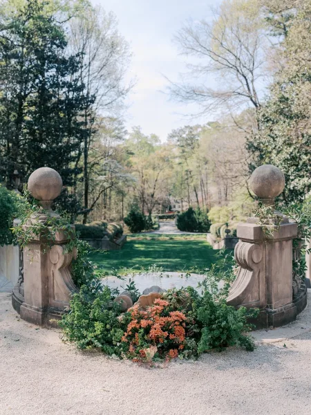Garden floral installation forming a ground floral arrangement flanking stone pillars by a gravel path and stone steps among hedges.